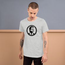 Load image into Gallery viewer, Man standing against a wall, wearing a heather grey unisex t-shirt, featuring the Cuomo Sapiens S.U.D.L.T. SEAL graphic in black.