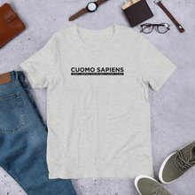 Load image into Gallery viewer, A heather grey unisex t-shirt on the floor, featuring the Cuomo Sapiens S.U.D.L.T. graphic in black.