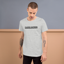 Load image into Gallery viewer, Man standing against a wall, wearing a heather grey unisex t-shirt, featuring the Cuomo Sapiens S.U.D.L.T. graphic in black.