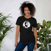 Load image into Gallery viewer, Woman standing next to some plants, wearing a black unisex t-shirt, featuring the Cuomo Sapiens S.U.D.L.T. SEAL graphic in white.
