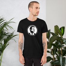 Load image into Gallery viewer, Man standing next to some plants, wearing a black unisex t-shirt, featuring the Cuomo Sapiens S.U.D.L.T. SEAL graphic in white.
