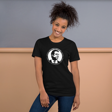 Load image into Gallery viewer, Woman standing against a wall, wearing a black unisex t-shirt, featuring the Cuomo Sapiens S.U.D.L.T. SEAL graphic in white.
