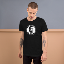 Load image into Gallery viewer, Man standing against a wall, wearing a black unisex t-shirt, featuring the Cuomo Sapiens S.U.D.L.T. SEAL graphic in white.
