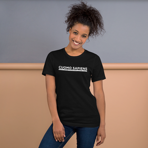 Woman standing against a wall, wearing a black unisex t-shirt, featuring the Cuomo Sapiens S.U.D.L.T. graphic in white.