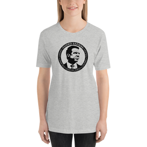 Woman wearing a heather grey unisex t-shirt, featuring the Cuomo Sapiens S.U.D.L.T. SEAL graphic in black.