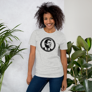 Woman standing next to some plants, wearing a heather grey unisex t-shirt, featuring the Cuomo Sapiens S.U.D.L.T. SEAL graphic in black.