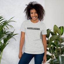 Load image into Gallery viewer, Woman standing next to some plants, wearing a heather grey unisex t-shirt, featuring the Cuomo Sapiens S.U.D.L.T. graphic in black.

