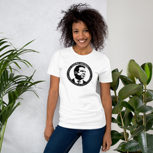 Load image into Gallery viewer, Woman standing next to some plants, wearing a white unisex t-shirt, featuring the Cuomo Sapiens S.U.D.L.T. SEAL graphic in black.
