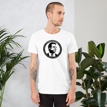 Load image into Gallery viewer, Man standing next to some plants, wearing a white unisex t-shirt, featuring the Cuomo Sapiens S.U.D.L.T. SEAL graphic in black.
