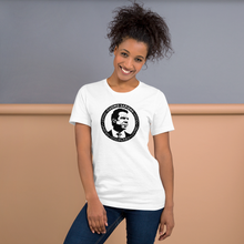 Load image into Gallery viewer, Woman standing against a wall, wearing a white unisex t-shirt, featuring the Cuomo Sapiens S.U.D.L.T. SEAL graphic in black.

