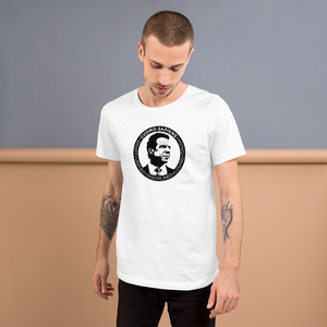 Man standing against a wall, wearing a white unisex t-shirt, featuring the Cuomo Sapiens S.U.D.L.T. SEAL graphic in black.